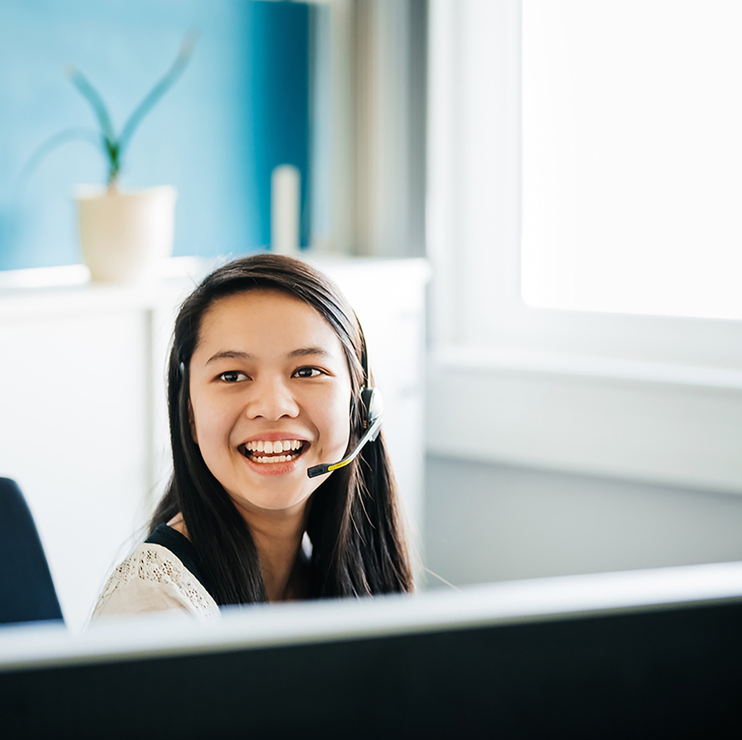 An office employee wearing a hands free headset laughing while sitting at her desk and working