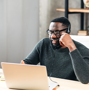Clever young African-American guy wearing stylish eyeglasses with headset sits on his workplace in the modern office, he looks at laptop screen, smiles and talking online