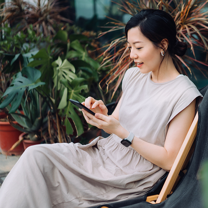 Beautiful young Asian woman using smartphone while relaxing on deck chair in the backyard, surrounded by beautiful houseplants